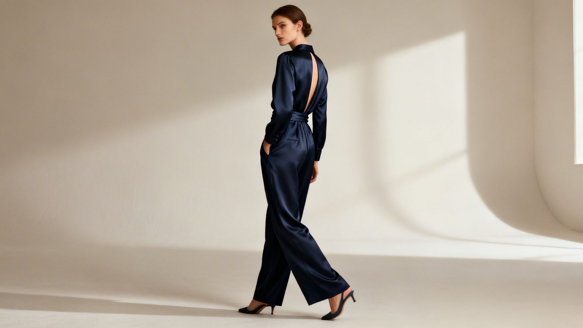 Kobieta w eleganckim, granatowym jedwabnym kombinezonie z odkrytymi plecami, idąca i patrząca przez ramię.
