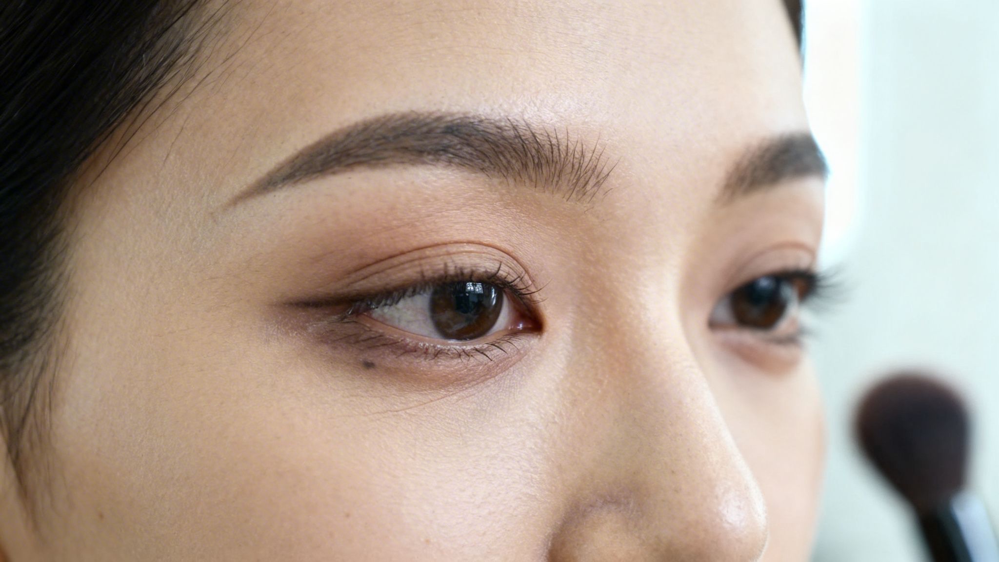 Zbliżenie na oko kobiety z brązowymi oczami, precyzyjnym makijażem i zadbanymi brwiami, w tle pędzel.