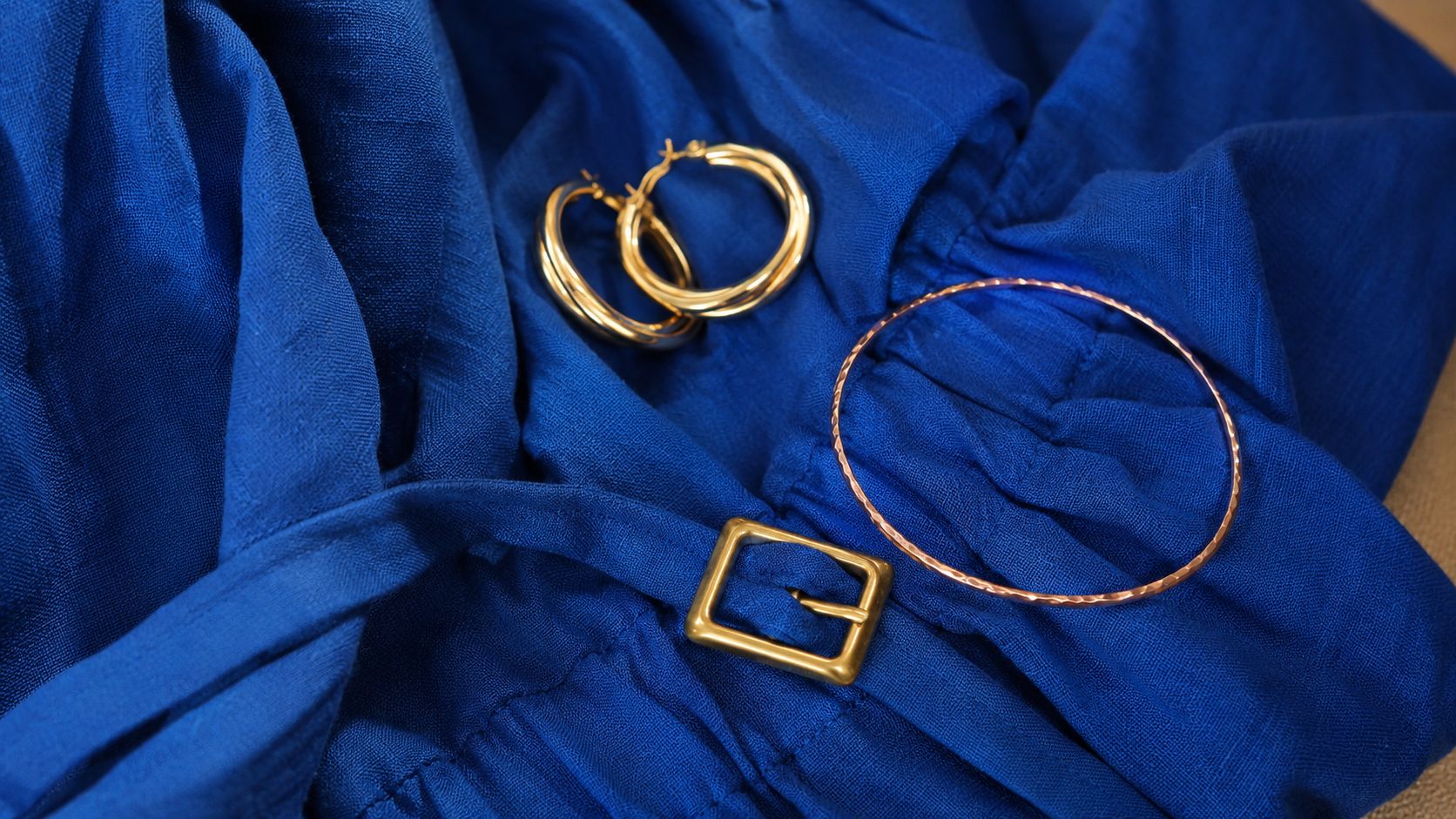 Złota biżuteria, kolczyki koła i bransoletka leżące na eleganckim, niebieskim materiale z ozdobną klamrą paska.