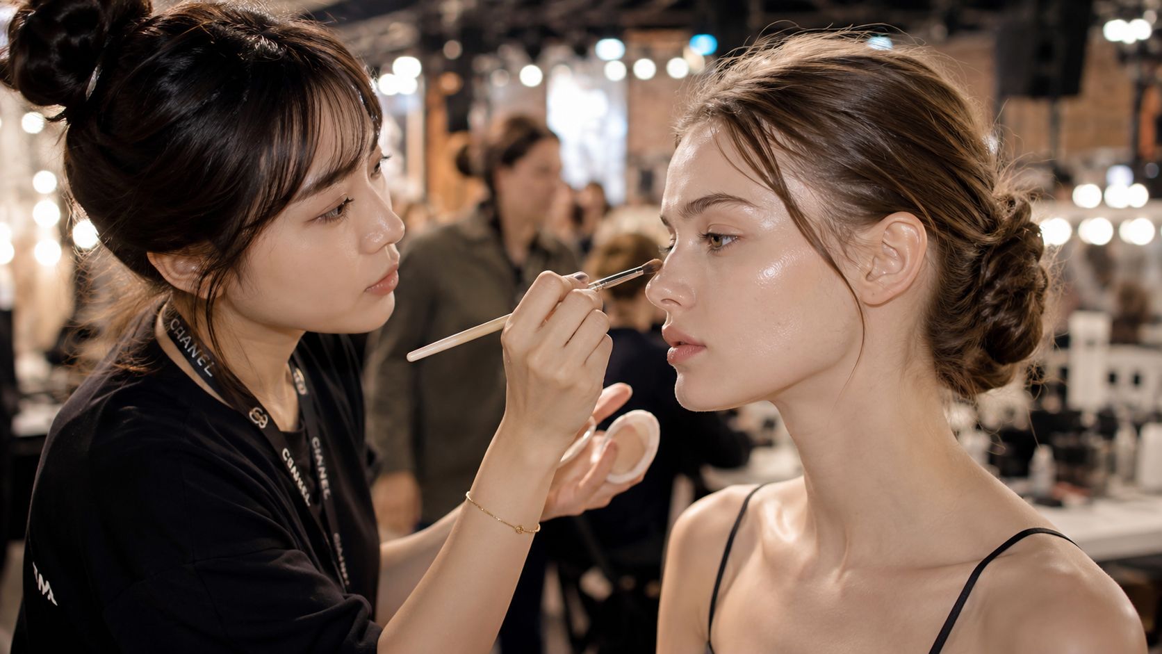 Makijażystka z logo Chanel nakłada profesjonalnym pędzlem rozświetlający makijaż na twarz młodej modelki w profesjonalnym studio.